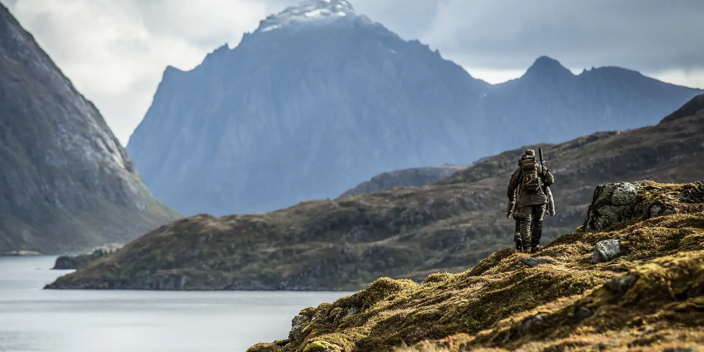 A hunter walks a mossy ridge above a Lofoten fjord, rifle slung and pack on, with the mountains ahead.